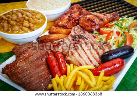 Platter of assorted grilled and barbecued meats, such as beef, pork, chicken and sausages, accompanied by brown beans, salad, fried plantains, french fries and white rice. Royalty-Free Stock Photo #1860068368