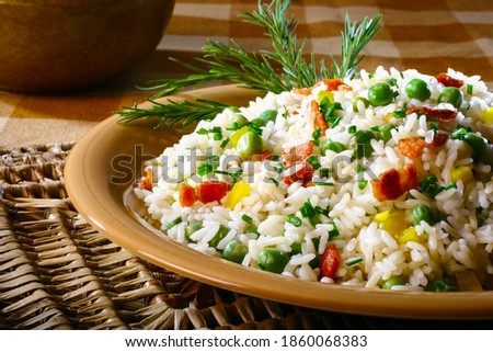 A dish of stuffed white rice with chives, green peas, corn and bacon pieces, all on a ceramic plate over a place mat on a table. Royalty-Free Stock Photo #1860068383