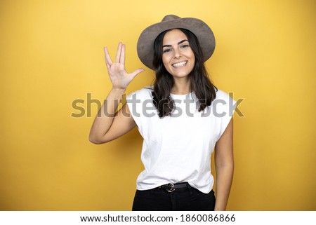 Beautiful woman wearing casual white t-shirt and a hat standing over yellow background doing hand symbol