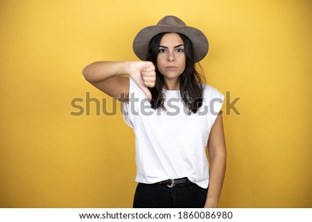 Beautiful woman wearing casual white t-shirt and a hat standing over yellow background with angry face, negative sign showing dislike with thumb down