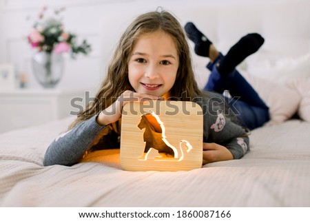 People, children, domestic life concept. Wooden home decorations. Adorable little pretty girl lying on the bed in cozy light room and playing with wooden night lamp with lion cut out picture