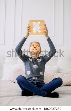 Indoor portrait of pretty school girl in casual domestic wear, sitting on the bed in lotus position and holding stylish wooden night lamp with bear picture under her head.