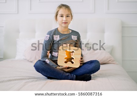 Indoor portrait of pretty school girl in casual domestic wear, sitting on the bed in lotus position and holding stylish wooden night lamp with bear picture.