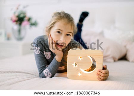 Little pretty 10 years old girl, lying on the bed at cozy light bedroom at home, and posing to camera with wooden stylish night lamp with cut out moon and stars picture. Wooden decorations
