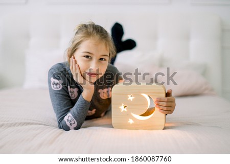 Little pretty 10 years old girl, lying on the bed at cozy light bedroom at home, and posing to camera with wooden stylish night lamp with cut out moon and stars picture. Wooden decorations