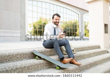 Happy businessman taking a break from work outside his office and texting on his phone. Male active worker with a skateboard