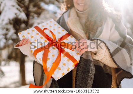Big gift box with red ribbon in the woman hands.  Fashion young woman holding a Christmas present standing among snowy trees and enjoying first snow. Holidays, season and leisure concept. 