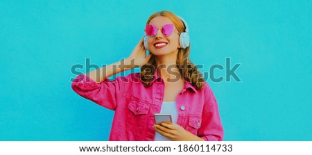 Portrait of stylish smiling young woman in wireless headphones with phone listening to music wearing a pink jacket, sunglasses over blue background