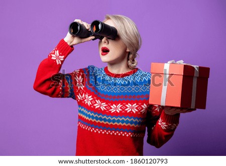 Beautiful woman in Christmas sweater with gift box and binocular on purple background