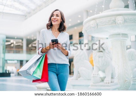 Attractive confident young woman standing in the mall with smartphone and a bunch of shoping bags in hands. Royalty-Free Stock Photo #1860166600