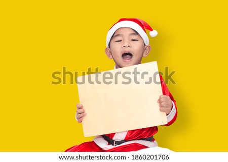 happy child wearing a santa claus costume with a wooden sign To enter text A picture of a boy with a mouth open and smiling against a yellow background. with clipping path.