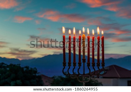 Glitter lights of candles on menorah are traditional symbols for Jewish Hanukkah Holiday of Light. Selective focus on candles. Background with blurred dramatic morning sky and mountains as background
