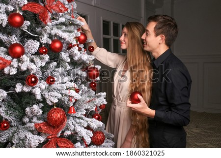long-haired young girl in a dress and a guy in a black shirt and trousers decorate a christmas tree with red balls and toys, festive home decor, new year atmosphere