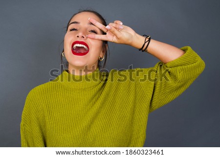 Young beautiful hispanic woman wearing green sweater against gray background Doing peace symbol with fingers over face, smiling cheerful showing victory
