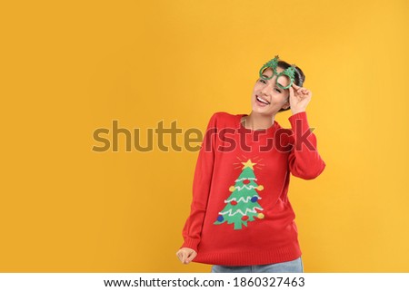 Young woman in Christmas sweater and party glasses on yellow background, space for text