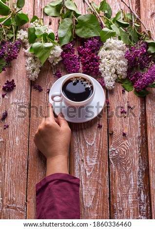 Hand holds a cup of morning coffee with spring lilac flowers branches blossoming on wooden background view from above. Flat lay underground style. Expensive colors. Creative design of flowers.