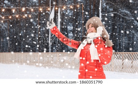 christmas, technology and holidays concept - happy smiling woman in winter fur hat with tablet pc computer over outdoor ice skating rink on background