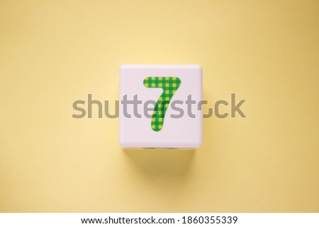 Close-up photo of a white plastic cube with a green number 7 on a yellow background. Object in the center of the photo