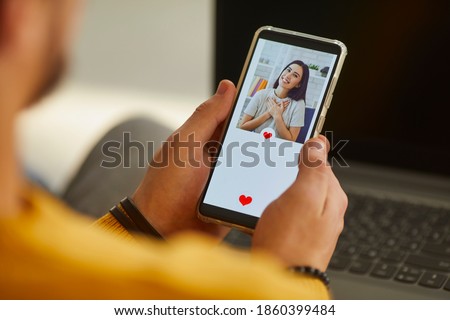 Find love online on social media or dating app concept. Over shoulder close-up single man looking at mobile phone screen and pressing red heart like button below attractive young woman's profile photo