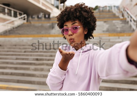 Beautiful pretty curly brunette dark-skinned woman in stylish pink hoodie and colorful sunglasses blows kiss and takes selfie near stairs outside.