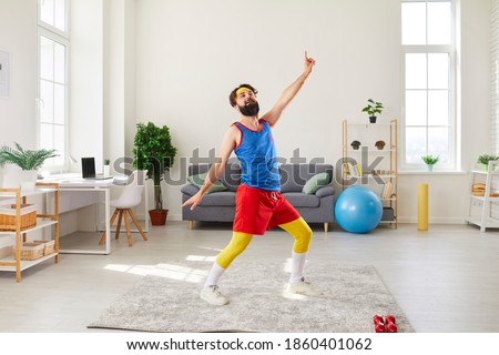 Funny bearded young man in colorful retro sportswear doing fitness exercise, dancing and fooling around in his living-room during sport workout at home
