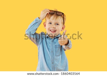 Portrait of surprised cute little child boy in sunglasses. Child having fun isolated over yellow background. Looking at camera. Showing thumbs up sign Ok. Wow funny face. 