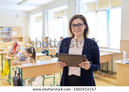 Portrait of mature woman teacher in classroom with digital tablet, smiling confident female on background of classroom with teenage students