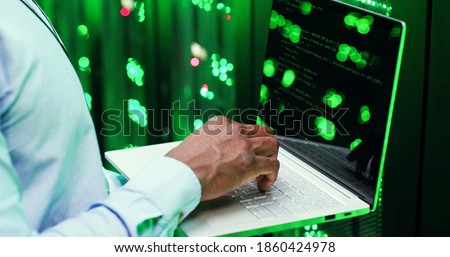 Close up of African American man among servers working on laptop computer, typing on keyboard and decrypting codes. Male analytic in data storage working on digital security and programming.