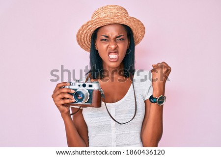 Young african american woman wearing summer hat holding vintage camera angry and mad raising fist frustrated and furious while shouting with anger. rage and aggressive concept. 