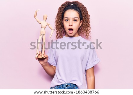 Beautiful kid girl with curly hair holding small wooden manikin scared and amazed with open mouth for surprise, disbelief face 