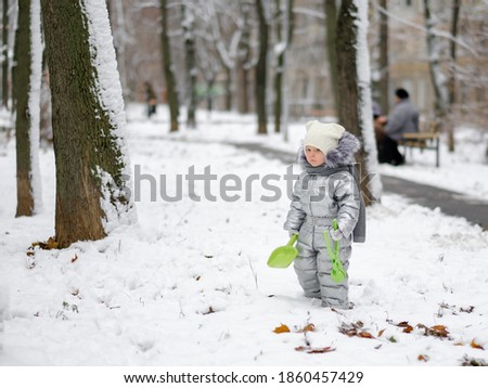 Happy child on a winter walk in the park. A funny little girl in a warm silver jumpsuit holds a toy shovel. First snowfall. Dry maple leaves peek out from under the snow. Blurred background.