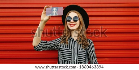 Portrait of happy smiling young woman taking selfie picture by smartphone wearing a black round hat, striped shirt over red background