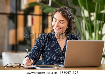 Successful young business woman working on laptop with headphones in a call center as a consultant. Happy businesswoman with headset translating and writing notes by listening to audio course. Royalty-Free Stock Photo #1860471397