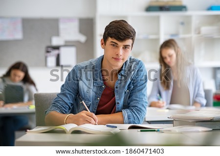 Happy student taking notes while studying in high school. Satisfied young man looking at camera while sitting at desk in classroom. Portrait of college guy writing while completing assignment. Royalty-Free Stock Photo #1860471403