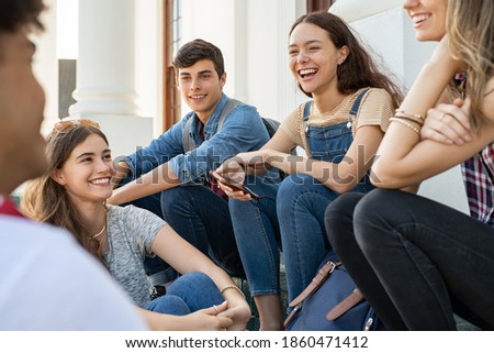 Group of happy young friends sitting in college campus and talking. Cheerful group of  smiling girls and guys feeling relaxed after university exam. Excited millenials laughing and having fun outdoor. Royalty-Free Stock Photo #1860471412