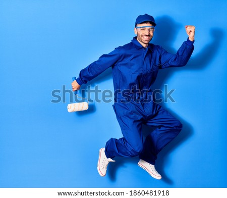 Young handsome hispanic man wearing painter uniform and cap smiling happy. Jumping with smile on face holding roller doing winner sign with fists up over isolated blue background