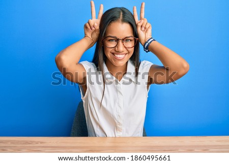 Beautiful hispanic woman wearing casual clothes sitting on the table posing funny and crazy with fingers on head as bunny ears, smiling cheerful 