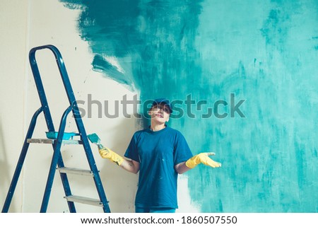 A young woman in work clothes tiredly wearily in perplexity throws up her hands while preparing the wall for painting