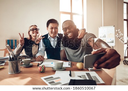 People showing victory sign and smiling for camera. Cheerful workers sit together at the table and smile while looking at the camera. 