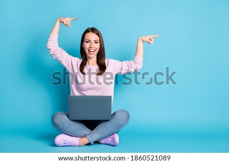 Full length photo positive girl sit floor legs crossed work remote laptop point index finger copyspace indicate repost ads wear lilac violet jumper denim jeans isolated blue color background