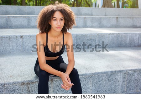 Picture of young African-American woman runner in a black trendy tracksuit on the stairs, resting after sports activities in the park. Sport, outdoor, healthy concept.