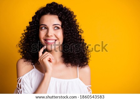 Photo portrait of interested young girl looking at empty space touching chin with finger smiling isolated on vivid yellow color background