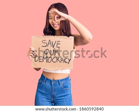 Young beautiful chinese girl holding save our democracy protest banner smiling happy doing ok sign with hand on eye looking through fingers 