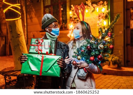 Close up portrait of joyful lovely mixed-races couple standing in evening in city with christmas presents in medical masks. Man and woman on snowy street looking at camera and smiling. Xmas concept.