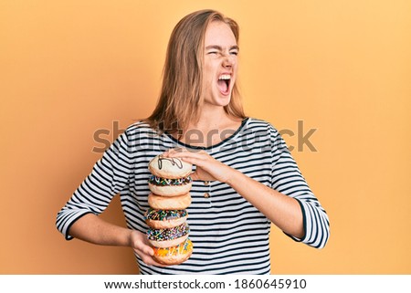 Beautiful blonde woman holding tasty colorful doughnuts angry and mad screaming frustrated and furious, shouting with anger. rage and aggressive concept. 