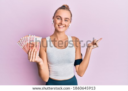 Beautiful blonde sport woman holding 100 norwegian krone banknotes smiling happy pointing with hand and finger to the side 