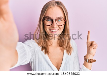 Beautiful blonde woman taking a selfie photo wearing glasses smiling with an idea or question pointing finger with happy face, number one 