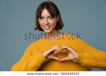 Young happy beautiful woman standing isolated over blue background, showing heart gesture