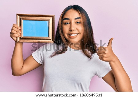 Young latin woman holding empty frame smiling happy and positive, thumb up doing excellent and approval sign 