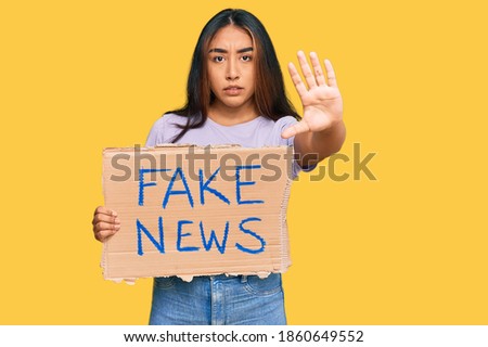 Young latin woman holding fake news banner with open hand doing stop sign with serious and confident expression, defense gesture 